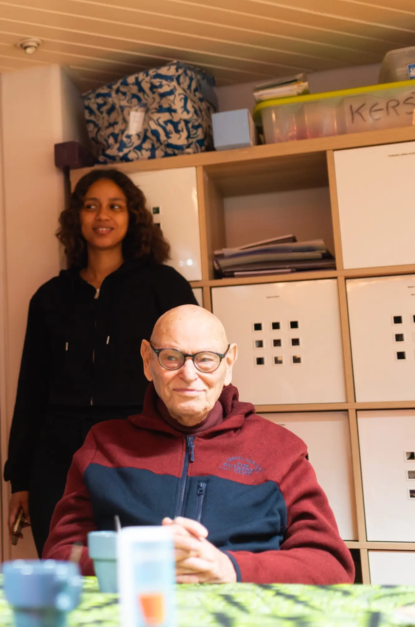 Een jonge vrijwilliger staat achter een oudere man die in een huiskamer-setting zit. Ze glimlachen samen, een mooi voorbeeld van hoe jongeren ouderen gezelschap houden en ondersteunen in het Boeken en Boterhammen-project.
