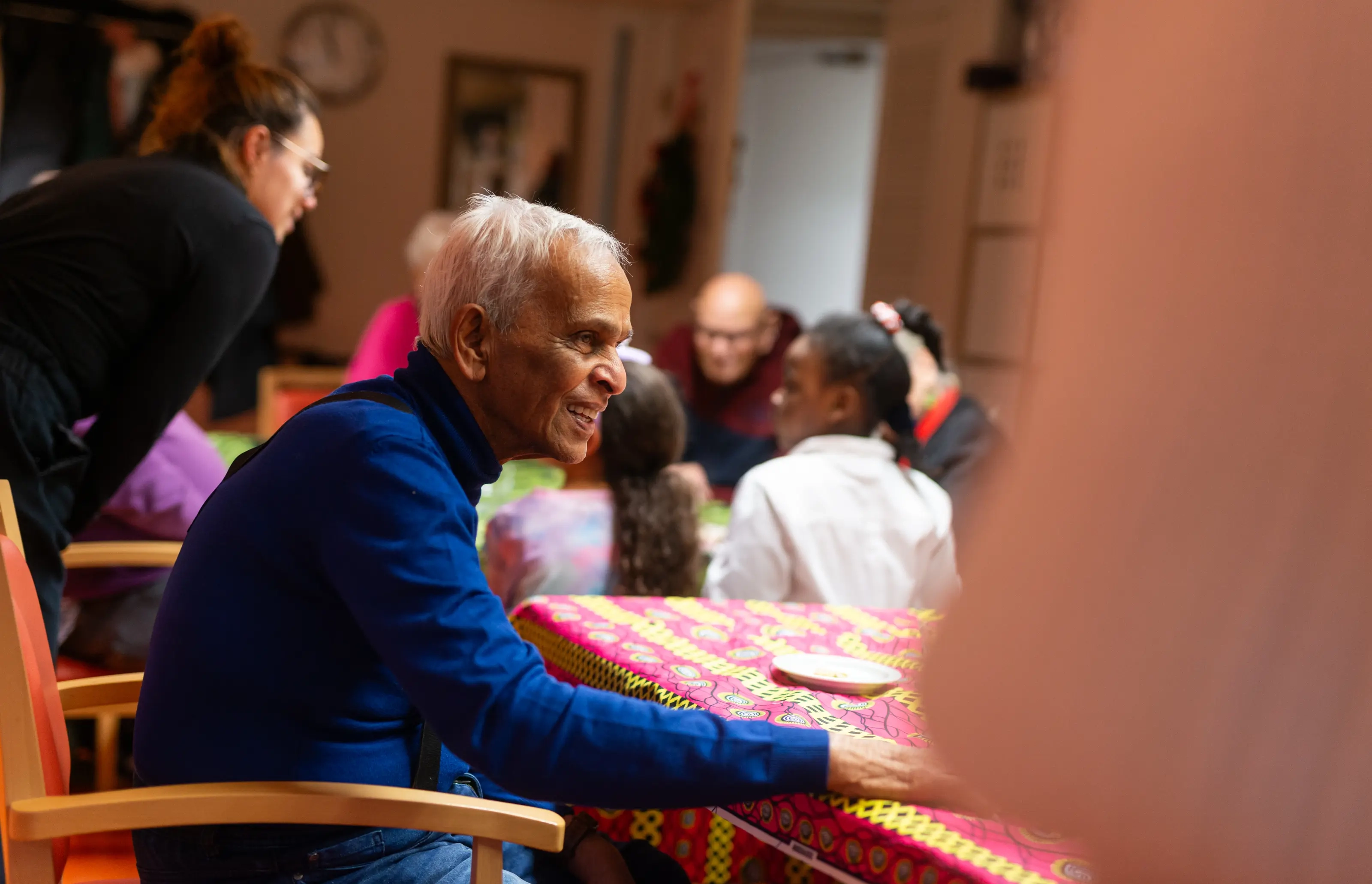 Een oudere man zit aan een tafel en kijkt naar een jonge vrouw die met hem in gesprek is. Dit laat zien hoe jongeren en ouderen binnen het project met elkaar in contact komen en samen een fijne tijd hebben.