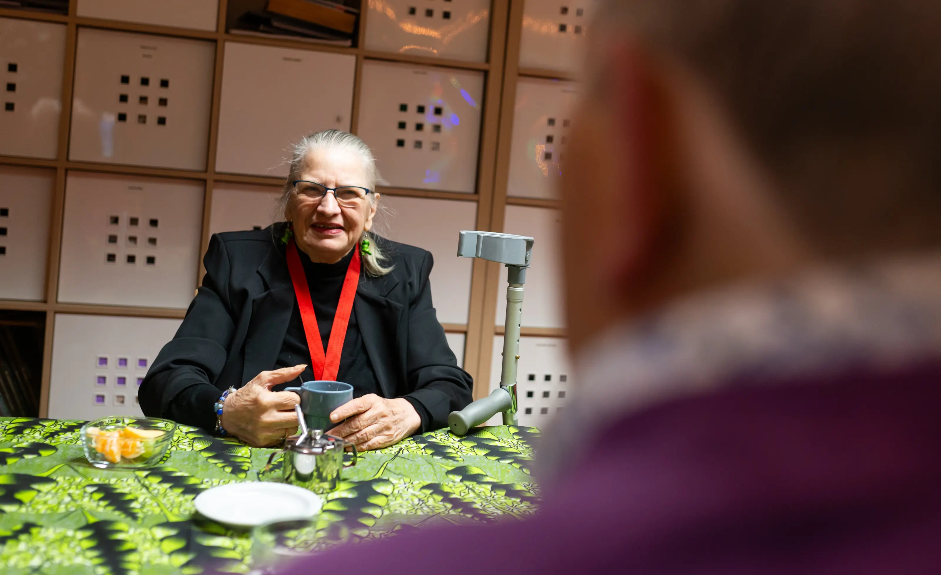 Een oudere man met een rode medaille om zijn nek zit met een kopje thee, terwijl een jongere naar hem luistert. Dit soort momenten maken het Boeken en Boterhammen-project bijzonder: jongeren en ouderen die elkaar echt leren kennen.